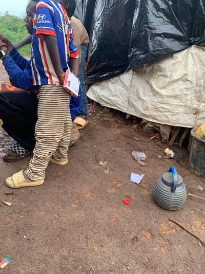 This image depicts a group of people in a makeshift shelter constructed from a black tarp and a white, weathered material. The ground is composed of damp, reddish-brown soil with scattered debris. Two individuals are prominently featured, seated on the ground. One person wears a blue and white striped long-sleeved shirt with red accents and the Hyundai logo on the sleeve. This person is holding a white and red box of medication with visible text. They are also wearing striped pants. Another person is visible behind them, wearing blue jeans.
In the background, a grey and blue ridged watering can is resting on the ground, next to scattered pieces of paper and what appear to be small red cylindrical objects. The overall scene suggests a humble dwelling or temporary shelter, possibly in a rural area given the natural ground and materials used for construction. The lighting suggests it is daytime. The image captures a moment of rest or interaction within this environment. The text on the medication box is partially visible and reads "Ziprolac capsule".