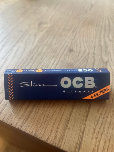 A pack of OCB Ultimate Slim rolling papers and filters lies on a wooden surface. The pack is navy blue with silver and orange accents. The text on the pack indicates it contains 32 papers and 32 filters. The surface beneath is a light brown wood. There is no location in the city of Ede, Netherlands in the image.
