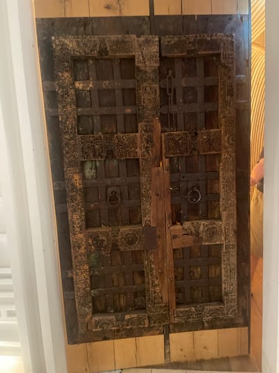 An ornate, dark wood, double door, featuring intricate carved detailing and metal reinforcements, stands slightly ajar. The door is set within a light-colored wooden frame and appears to be an interior entrance. A person, wearing a dark top and tan pants, is partially visible on the right side of the frame, appearing to walk away from the door. The background on the right shows a curved, textured surface, possibly a wall or partition, in a warm, light-brown hue. The left side of the image shows a white, vertical architectural element, likely a wall or pillar. The scene is indoors. The object is located at the Museum of Gukak in Gimpo-si, South Korea.