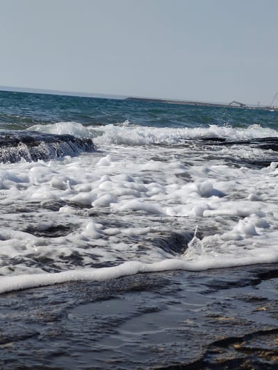 A low-angle, medium shot captures a churning sea with foamy white waves crashing against dark, wet rocks in the foreground. The water is a deep teal with lighter, frothy areas where the waves break. The sky above is a pale, clear blue. In the far distance, a long, dark breakwater stretches across the horizon, with industrial cranes and structures visible on it. The image focuses on the dynamic interaction of the water and the rocky shore, with no people or animals present. The scene evokes a sense of a natural coastal environment, possibly near a port or industrial area.