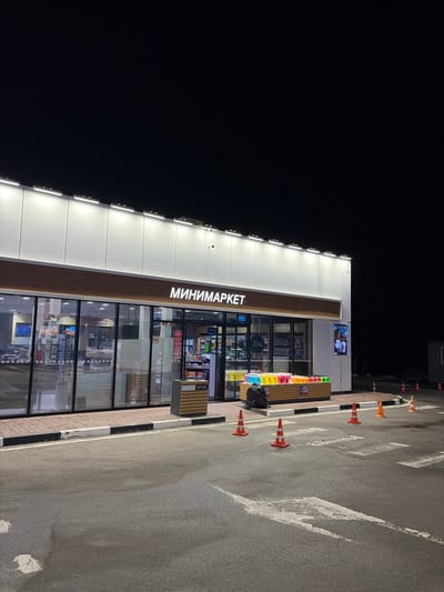 A brightly illuminated minimarket stands on an asphalt surface at night. The storefront is mostly glass, revealing shelves of goods inside. Orange cones line the parking area. The building's facade is light-colored with the word "МИНИМАРКЕТ" displayed vertically in Cyrillic. A black garbage bag sits near a wooden bench outside the store. The sky is dark.