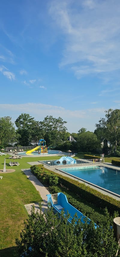 This is an aerial, full shot of a brightly lit outdoor swimming complex on a clear day. The complex is situated in Groenlo, Netherlands, as indicated by the specific detail. The foreground features a blue water slide and lush green trees. In the mid-ground, a well-maintained lawn is dotted with lounge chairs and umbrellas, leading to a wading pool with a yellow slide and colorful playground structure. Further back, a large rectangular swimming pool, partially covered by a blue inflatable structure, is visible. The background consists of a line of trees and houses, with a clear blue sky overhead. There are no people visible in the image.