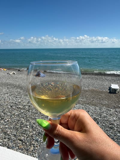 A hand with bright green painted nails holds a wine glass filled with pale yellow liquid. The glass is positioned in the foreground, with its stem and base extending out of frame to the right. The wine reflects light, creating prismatic effects within the glass.
The background depicts a natural outdoor scene, likely a beach. The left side of the image shows a clear blue sky with scattered white clouds. To the right of the sky is the ocean, a vibrant teal color, with gentle waves lapping at the shore. The beach itself is covered in dark gray pebbles. In the middle ground, a white, rectangular object, possibly a small structure or piece of furniture, is visible on the pebble beach.
The overall atmosphere suggests a leisurely day, perhaps a vacation, at the seaside. The bright sunshine and the presence of a beverage imply a relaxed and enjoyable time. The specific location is identified as Tsandrypsh, Abkhazia. The time of day appears to be daytime, likely late morning or afternoon, given the bright sunlight casting shadows. No visible text is present in the image.
