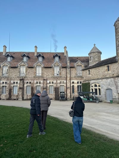 The image displays an outdoor scene at a large stone building in Cny-la-Ville, France, under a clear blue sky. Three individuals walk from left to right in the foreground. The leftmost person, with their back mostly to the camera, wears a dark puffy jacket with a white logo, dark pants, and a grey beanie, with their left leg extended. The central person, partially obscured, wears a light brown or grey puffy jacket and dark trousers. The person on the right, with their back to the camera, wears a dark hooded jacket, blue flared jeans, and carries a dark handbag over their left shoulder. They are situated on a grassy lawn adjacent to a light-colored dirt path.
The background is dominated by a multi-story stone building featuring a brown tiled roof with multiple dormer windows and several chimneys. A thin column of grey smoke rises vertically from one central chimney. A prominent circular stone tower with a conical, cross-topped roof is visible on the right side of the building, with a taller, narrower tower partially visible on the far right edge. Bare vines cling to sections of the building's stone facade. Two utility vehicles are parked on the dirt path: a dark, open-topped cart with a canvas cover is positioned to the left of the green cart, and a green golf cart is parked to the right, near a building entrance. A black bicycle is partially visible behind the green golf cart, and a small green bin is near a grey door on the building's right side. The sky above is clear and bright blue.