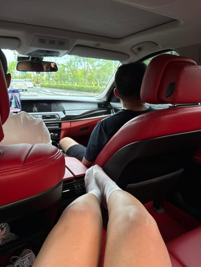 The image shows the interior of a car with red leather seats, as viewed from the back seat. Two individuals are visible: one person in the passenger seat, facing forward, and another person in the driver's seat, also facing forward. The person in the foreground has their legs extended, wearing white socks. Outside the car, a street scene with trees and other vehicles is visible through the windows. The car appears to be moving through the city of Lin'an District, China, suggested by the presence of cars and the overall urban environment.