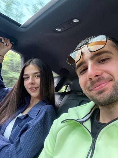 Two people are in a car, taking a selfie. The man is wearing glasses and a light green jacket, while the woman is wearing a blue pinstripe blazer. The location is unknown.