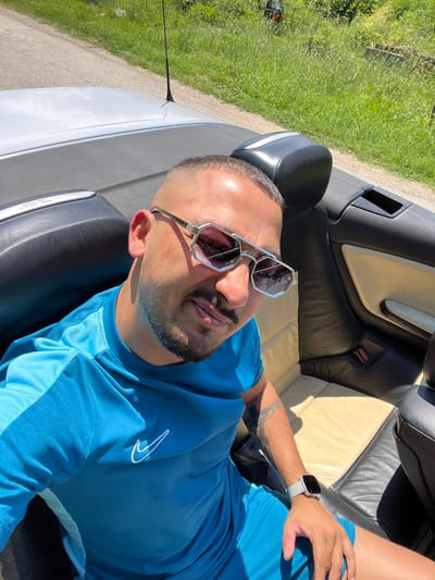 The image shows a man in a blue t-shirt and shorts sitting in the passenger seat of a convertible car. He is wearing sunglasses and a smartwatch. The car's top is down, and lush green vegetation and a road are visible in the background. The man appears to be in Batoshevo, Bulgaria, based on external information not present in the image itself.