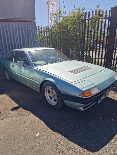 A light blue vintage Ferrari, likely a 400 or 412 model, is parked outdoors on an asphalt surface under clear, sunny daylight. The car is viewed from the front-right quarter, showing its angular design, pop-up headlights (retracted), and a rectangular louvered vent on the hood. The front bumper is dark, and the front right wheel, with a silver five-spoke design and a visible Ferrari prancing horse logo on the hubcap, is prominently featured.
Strong shadows are cast on the ground to the right of the car, indicating a high sun position. In the background, a dark brown metal fence runs horizontally, behind which dense green foliage of bushes or trees is visible. Further in the background, a light-colored commercial banner is partly visible against the blue sky, displaying the text "MICK ASH" and partially "SERVICES / STORAGE". Another segment of the banner reads "HIRING".
The vehicle bears a UK registration plate on its front, which appears to read "D 923 MXY", indicating a registration from late 1986. The setting suggests a commercial or light industrial area, consistent with the fence and business signage, within South Tyneside, United Kingdom. The image captures a static scene with no discernible activity or emotions.