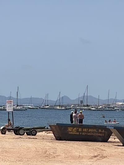 In the foreground of the image, a sandy beach area is visible, with tire tracks imprinted in the sand. A dark, large metal skip container with yellow lettering that reads "EFB HIJOS, S.I. ABANDOS Y EXCAVACIONES" is positioned on the beach, partially obscuring the view of the water. To the left of the skip, a two-wheeled trailer is parked on the sand.

In the middle ground, the sea is calm and blue. A group of four individuals are standing near the edge of the skip, facing towards the water. One individual is wearing a yellow hat. Further out in the water, several people are visible, some in small boats or on inflatable devices. A prominent sign with Spanish text is visible on a pole to the left, near the water's edge, with a person sitting on the ground nearby.

The background of the image features a harbor filled with numerous sailboats and yachts, their masts creating a distinct silhouette against the horizon. Beyond the harbor, a range of mountains can be seen under a clear, pale blue sky. The overall scene depicts a coastal area, likely in the city of San Pedro del Pinatar, Spain, characterized by a beach, a harbor with boats, and a mountainous backdrop.