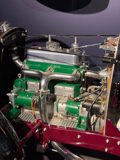 This image captures a meticulously preserved engine, likely from an antique vehicle, displayed indoors. The engine features prominent bright green painted components, including two main blocks or covers, which contrast with polished silver or chrome pipes and heat sinks.
On two of the green engine covers, the embossed text "LEA-FRANCIS COVENTRY" is clearly visible, identifying the manufacturer and place of origin. Intricate wiring, including red, orange, yellow, blue, and black insulated cables, connects various electrical components on the engine. Numerous bolts, nuts, and metal fasteners are visible throughout the assembly, along with several intricate mechanical parts and tubes.
To the right, a glossy, dark red painted metal frame or chassis component runs vertically, secured by silver bolts, suggesting part of the vehicle's structure. Portions of what appears to be a dark tire or wheel and reflective chrome bodywork are visible at the bottom.
The setting appears to be a museum or exhibition space, with a plain, dark purple-grey background and artificial lighting that highlights the engine's details. No activity is taking place; the engine is stationary and presented for observation. The scene is located in Wassenaar, Netherlands.