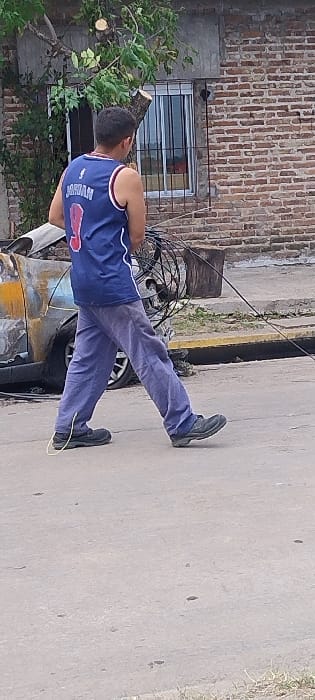 An outdoor scene captured in San Fernando, Argentina, features a man standing near a damaged vehicle on what appears to be a concrete or paved surface. The image is taken during the daytime, under clear weather conditions.

The main subject is a man, seen from a slightly elevated back and side angle. He is wearing a blue sleeveless jersey with white and red accents, clearly displaying the name "JORDAN" and the number "23" on the back. His trousers are dark blue or grey, and he wears dark shoes. A thin yellow cable or rope is visible near his right foot, extending downwards.

To the man's right, a partially visible car appears old or damaged, with areas of yellowish-brown discoloration and dark, possibly burnt, marks. One of its wheels is visible. Nearby, a cluster of black bundled cables or wires lies on the ground.

In the background, on the left side of the frame, there is a brick building with a window protected by horizontal metal bars. Lush green foliage from bushes or trees is prominent on this side, with several cut tree branches, one showing a fresh circular cut mark. The ground is a uniform grey concrete or asphalt, with some minor imperfections.

The man's posture suggests he is standing still, possibly observing or engaged in an activity related to the surrounding elements, such as the car or cables. The presence of cut tree branches implies recent tree trimming in the vicinity.