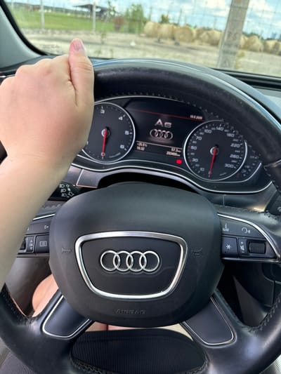 This media file shows a close-up view of a person's hand on the steering wheel of an Audi car. The steering wheel has the Audi logo in the center and controls on the left and right sides, including buttons for navigation and volume. The dashboard is visible, showing a speedometer, odometer, and a digital display with information such as temperature (-25.5°C), time (15:32), date (19.6.2026), fuel efficiency (57.3), and mileage (293400). The car appears to be parked or stopped, as indicated by the static position of the hand and the dashboard readings. Through the windshield, a blurred outdoor scene with what looks like construction materials or hay bales and a clear sky can be seen, suggesting it is daytime with good weather. The overall impression is that of someone inside a car, possibly taking a break or preparing to drive. The visible text includes the car's model "A6" and various dashboard indicators. The location context provided is Kostiantynivka, Ukraine.
