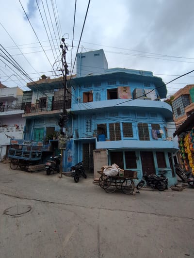 The image shows a street scene in Jodhpur, India, with a distinctive light blue building dominating the frame. The building has multiple stories, balconies with intricate metal railings, and a curved architectural element on the upper floor. Numerous electrical wires crisscross overhead, strung between a utility pole and various structures.
In front of the building, a blue cart with wooden sides and spoked wheels is parked. It appears to be a type of handcart or pushcart, possibly used for transporting goods. Several motorcycles are parked on the paved street, including two dark-colored scooters and a motorcycle with a rider in dark clothing.
The street is paved with concrete or asphalt and shows some signs of wear, with a visible crack running across it. A round metal manhole cover is also present on the street. The buildings on either side of the blue structure appear to be of similar architecture, with ornate windows and balconies. Some laundry is visible hanging from a balcony of the blue building. Bags of what appear to be groceries or merchandise are stacked on a wooden cart in the foreground. The sky is overcast with grey clouds.