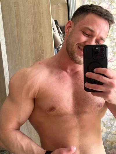 A shirtless man is taking a selfie with a black smartphone. He is holding the phone in front of his face. His face is partially obscured by the phone, but his smile is visible. He is fair-skinned with short brown hair and a beard. His chest and arms are visible, showing muscle definition. A wooden drawer is visible in the foreground. A floral patterned wallpaper is visible in the background. The location is Dobryanka, Russia.