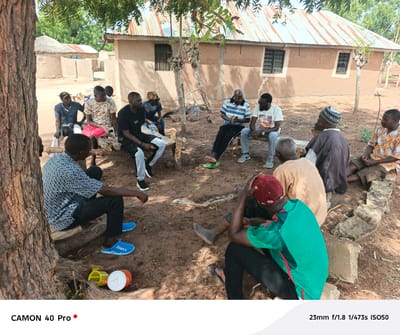 In Gidan Kogi, Nigeria, a group of men are gathered outdoors, seated in a circle. Some sit on wooden benches, while others are positioned on a low stone wall. The setting is a dirt clearing with a tree in the foreground. Buildings with corrugated metal roofs and mud walls are visible in the background. The men are of varying ages and are dressed in casual clothing. Some are engaged in conversation, while others are listening. One man is barefoot. Several plastic containers are near the base of the tree.