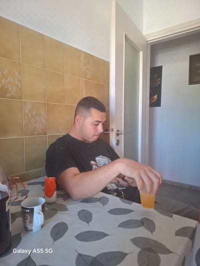 The image depicts an indoor residential setting, identified as being within the Heraklion Municipal Unit, Greece. One light-skinned adult male with short, dark hair is seated at a table, looking downwards. He is wearing a dark-colored t-shirt with a faint graphic pattern resembling currency. His right hand holds a smartphone oriented downwards, and his left hand is positioned over a transparent plastic cup containing an orange liquid.
The table is covered with a light-colored tablecloth adorned with a pattern of grey leaf silhouettes. On the table, from left to right, are a partially visible dark bottle, a copper-colored wire stand, a white ceramic mug inscribed with "CAPPUCCINO" and coffee-related graphics, and an orange bear-shaped condiment dispenser with a white base.
The background consists of a wall tiled with light brown square tiles, many featuring a repeating decorative pattern of white, stylized wheat or grass stalks. Above the tiled section, the wall transitions to a plain white ceiling. To the right of the seated male, a white door frame and an ajar white door with silver hardware provide a view into an adjacent room. This room has light blue-grey walls and displays two framed artworks: a dark-toned piece with an orange stylized floral design on the left, and a less distinct framed image on the right. A dark skirting board runs along the base of the wall in the adjacent room. The scene is illuminated by ambient light. A "Galaxy A55 5G" watermark is visible in the bottom-left corner of the image.