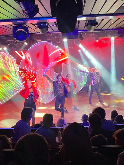The image depicts a performance on a brightly lit stage, with performers in costumes and a large LED screen displaying abstract patterns. The lighting is dynamic, with spotlights creating beams of light. The audience is visible in the foreground, seated in what appears to be a theater or concert hall. The performers are engaged in some form of dance or theatrical routine. The overall atmosphere is energetic and visually engaging. The location is specified as Hitis, Finland, though no specific landmarks or context are provided within the image to confirm this.