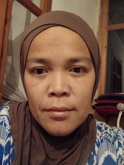 A medium shot shows a woman wearing a brown hijab and a blue and white patterned shirt, looking directly at the camera. Her face fills most of the frame. Her skin tone is medium brown. She has dark eyes, a small nose, and her lips are slightly parted. The background is dimly lit and appears to be indoors, with wooden elements and a textured wall visible. To the right, a stack of reddish-purple cushions is partially visible. The setting is indistinct, but the overall impression is of a simple, personal space. There is no indication of the city of Kotormo, Kyrgyzstan.
