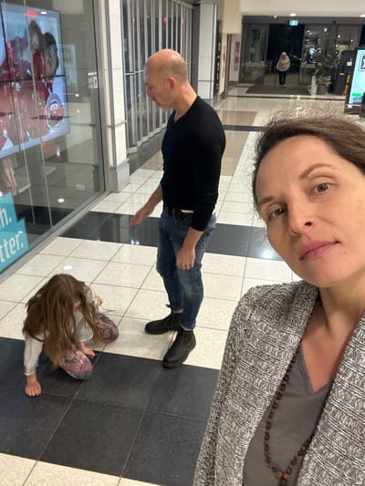 In Toormina, Australia, within a shopping center hallway, a bald man in a black long-sleeved shirt and blue jeans stands looking at a large display screen. To his left, a young girl with long brown hair is kneeling on the floor, seemingly playing or examining something. In the foreground, a woman with brown hair and a grey patterned cardigan is taking a selfie, her face angled towards the camera. The hallway features a tiled floor with contrasting black and light-colored sections, glass storefronts, and automatic doors in the background leading to an exit.