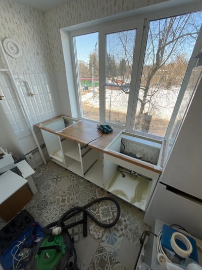 The image displays an interior room, likely a kitchen undergoing renovation, located in Нарынка, Russia. The perspective is directed towards a large window, with the floor and ongoing construction elements in the foreground.
The left wall features light-colored wallpaper patterned with a subtle botanical design of leaves and stems. Below the wallpaper, a section of white subway tiles extends across the wall. White heating pipes, including a radiator with a circular white cover, are mounted on this tiled section, along with two brass-colored spigots.
A wide, white-framed window, composed of multiple panes, dominates the back wall. It provides a view of an outdoor environment characterized by patches of snow on the ground, numerous bare deciduous trees, and some distant evergreen trees, indicative of winter or early spring. A building with a red roof and other structures are visible in the middle distance under a clear, bright sky.
Directly beneath the window, white base cabinet frames are partially installed. A light-brown wooden countertop is fitted over a central section of these cabinets. A teal-colored power tool, likely a jigsaw, with a black electrical cord, rests on this wooden surface. Two rectangular cutouts, intended for sinks or appliances, are visible in the counter assembly. The rightmost cutout reveals plumbing pipes within the cabinet below, whose base is lined with a yellowish waterproof material. Open white cabinet compartments are visible below the wooden countertop.
The floor is covered with ceramic tiles featuring a complex, multi-patterned design in shades of grey, blue, and brown.
In the lower left foreground, a black vacuum cleaner hose is coiled on the floor, adjacent to an open dark-colored tool bag containing various hand tools, including pliers and screwdrivers, and a green object. In the lower right foreground, a section of a white refrigerator is visible. Beside it, on a lower surface, a blue item holds a roll of white masking tape, coiled electrical cords, and a white European-style electrical plug. No individuals are present in the scene.