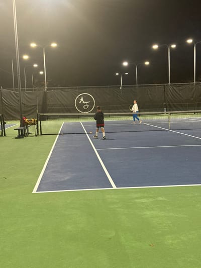 A tennis court at night is illuminated by overhead lights. Two individuals are present, one closer to the net, dressed in black and red, with tennis balls near them. The other person is further back, closer to the fence, wearing white and blue attire, holding a racket. A tennis ball basket sits on the court's edge. The court is a mix of blue and green surfaces.
