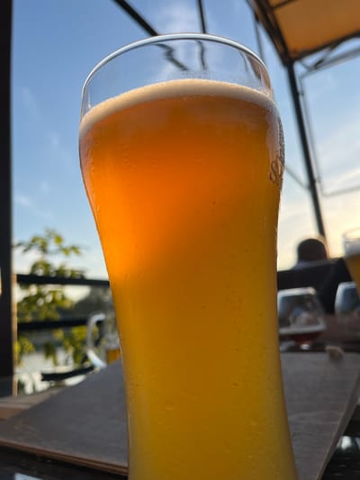This media file is a close-up, low-angle shot of a glass of beer. The beer is a bright, translucent yellow-orange color with a frothy white head. The glass is tall and curved, with condensation visible on the exterior. The background is blurred, showing a bright blue sky with some clouds and greenery, suggesting an outdoor setting during the day. The lighting indicates it is likely late afternoon or early evening, with the sun casting a warm glow. There are some faint markings on the glass, possibly a logo or brand name, including what appears to be a cursive "S". The overall mood is relaxed and casual, hinting at a pleasant outdoor gathering or leisure time. The location is specified as Azov, Russia. There are no discernible people in focus, only blurry figures in the background.