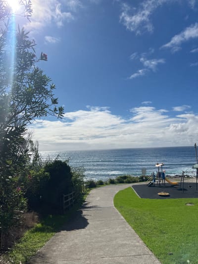 This image depicts a coastal public area in Coalcliff, Australia, under a bright blue sky with scattered white clouds and visible sunlight originating from the upper left, causing a lens flare. A light grey concrete pathway curves from the foreground, left to right, then towards the ocean. To the left of the pathway, dense green vegetation, including a tree with green leaves and pink flowers, and a low wooden fence are visible. To the right of the pathway, a patch of maintained green grass abuts a children's playground. The playground features a multi-colored play structure with a yellow slide and blue stairs, a circular spinning disc, and a partial view of a swing set, all situated on a black rubber soft fall surface. A white signpost is located near the play structure. The Tasman Sea occupies the background, displaying moderate waves and reflective sunlight on its surface. The coastline appears rocky or structured near the water's edge below the playground. No individuals are present within the scene.