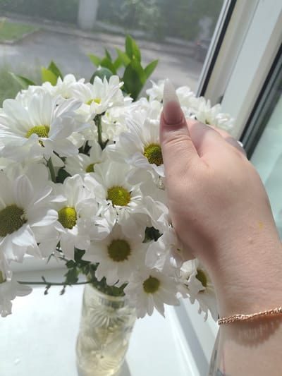 A close-up, slightly overexposed shot shows a bouquet of white daisies with bright green centers, arranged in a clear, patterned glass vase. A person's right hand, with a long, pointed, clear acrylic nail, is gently touching some of the flowers. The background reveals a window with a blurred view of trees and a street, suggesting an urban setting. A thin, rose-gold chain bracelet is visible on the person's wrist, and a tattoo can be partially seen on their forearm. The lighting suggests it is daytime, with sunlight streaming through the window. The image does not contain information that would indicate the specific location within Pervouralsk, Russia.