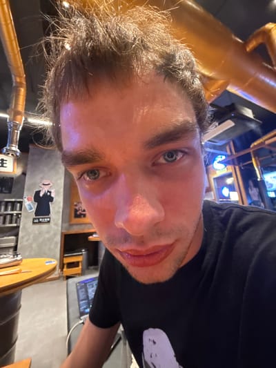 This media file is a close-up selfie of a man, rotated counter-clockwise, taken indoors in what appears to be a bar, brewery, or themed restaurant.

**Subjects:** The primary subject is a man, likely in his 20s or 30s, with short, dark, slightly disheveled hair and distinctive light blue-grey eyes, gazing directly at the camera. He has light stubble and his mouth is slightly open, giving him a neutral to slightly amiable expression. He is wearing a dark, likely black, t-shirt.

**Scene and Setting:** The background suggests an industrial or steampunk-inspired decor, featuring prominent golden or copper-colored pipes and possibly a tap in the foreground. The walls are dark grey, possibly concrete. The lighting is dim, creating an evening or nighttime ambiance, enhanced by blue and white artificial lights from screens or fixtures in the blurred background. Wooden tabletops or counters are partially visible.

**Activity or Event:** The man is taking a selfie, indicating a casual personal moment within a social venue.

**Notable Details:** A notable decorative element is a black and white graphic cutout on the wall, portraying a stylized figure resembling a "secret agent" or "mafioso" in a hat and trench coat, holding a briefcase. The specific location in Minhang District, China, aligns with the potential for such themed establishments.

**Visible Text:** Faint white text is visible on the briefcase of the graphic figure, and more text, likely Chinese characters, appears on the wall below the figure. A partial white label with the letter "E" is discernible on a wooden surface in the lower left. Additional indistinct text, possibly menu items, can be seen on a screen in the distant background.