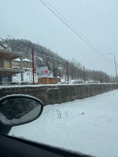 The image depicts a winter scene during a snowfall, viewed from inside a vehicle in the Raska Municipality, Serbia. The foreground shows a dark-colored vehicle interior on the lower left, including a snow-covered side-view mirror reflecting a person holding a phone. The immediate ground outside the vehicle is covered in a thick layer of fresh snow, with sparse, thin vegetation protruding.

A concrete retaining wall, approximately 1.5 meters high and covered in snow, extends horizontally across the mid-ground. Behind this wall, a chain-link fence supported by red-painted metal posts runs parallel to the wall. Numerous passenger vehicles, largely covered by snow, are parked behind the fence on a snow-covered surface.

To the left, behind the fence, two multi-story structures, possibly under construction or renovation with exposed concrete and some yellow and blue elements, are visible on a higher elevation. Adjacent to these structures and behind the fence, a white billboard features the "FG" logo and text in Serbian Cyrillic, "TEHNIČKI PREGLED" (Technical Inspection). Below the billboard and to its right, two rectangular wooden containers are present.

The background is dominated by a steep, snow-covered hillside densely forested with coniferous trees, all laden with snow. The sky is uniformly overcast and light grey, with visible snowflakes falling. Overhead power or communication lines traverse the upper portion of the image. On the far right, a snow-dusted street light pole with an extended arm is visible. No individuals are explicitly seen apart from the reflection in the side mirror.