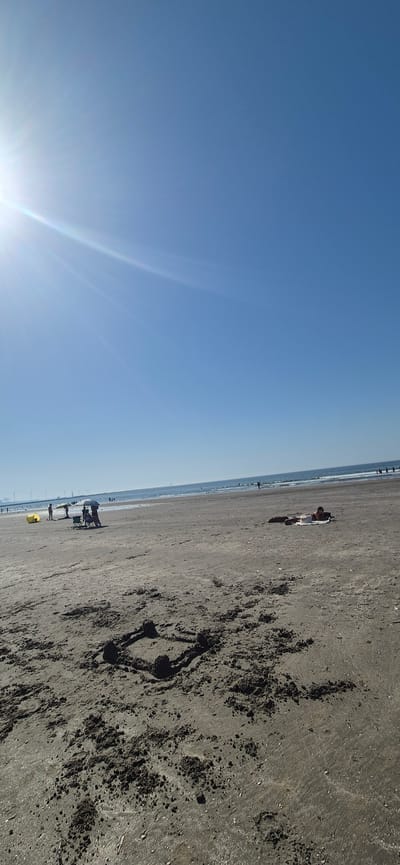 This is a photograph of Hook of Holland beach, Netherlands, taken on a clear, sunny day. The image captures a wide expanse of sandy beach with the calm sea in the background. The sun's rays are visible in the upper left corner, creating a lens flare effect. In the foreground, a sandcastle, roughly square in shape with four mounds at the corners, is present. Further down the beach, several people are visible. To the left, a group of individuals is gathered under a beach umbrella, with a yellow inflatable object nearby. Closer to the water, another person is seen lying on a towel, possibly sunbathing. The horizon shows a line of what appears to be wind turbines in the distance. The overall scene depicts a typical day at the beach with leisure activities taking place.