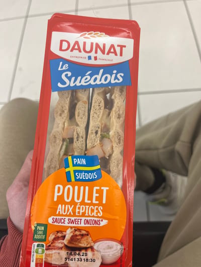 The image shows a hand holding a plastic-wrapped sandwich. The sandwich is labeled "Dauunat Le Suédois Poulet Aux Epices Sauce Sweet Onions." The label also displays a Swedish flag and the date 14.04.25 5141 33 18:30. The sandwich contains chicken and vegetables. The hand is wearing a brown shirt, and the background is blurred. The location is Saint-Brice-Courcelles, France, at latitude 49.283 and longitude 4. The scene is one of a person holding and likely consuming a packaged sandwich. The hand, sandwich, and packaging are the subjects. The environment is a public space. The action is the holding of the sandwich. The interaction is between the hand and the sandwich. The sandwich is not yet opened.