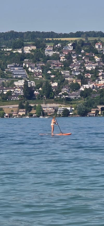 A lone individual is paddleboarding on a body of water, facing towards the left of the frame. They are wearing a light-colored bikini and a dark hat. The paddleboard is orange with a white stripe. In the background, the city of Oberrieden, Switzerland, is visible on a hillside. Numerous houses are clustered on the slope, surrounded by lush green trees. The sky is clear and blue.
