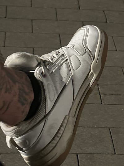 A close-up, low-angle shot captures a white leather sneaker with a gum sole on a person's foot. The shoe is worn, showing scuffs and dirt, particularly on the sole and toe box. The foot is clad in a dark sock, and a tattoo with bold, dark lines is visible on the ankle. The background consists of gray brick paving stones, casting distinct shadows, indicating the presence of sunlight. This image is not set in Siedlce, Poland; it is a generic urban outdoor setting.