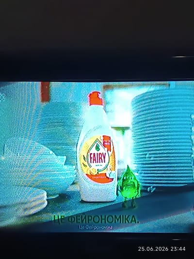 This is a photograph of a television screen displaying an advertisement. The advertisement features a bottle of "FAIRY" dish soap, a small green cartoon character, and stacked plates. Text at the bottom of the screen reads "ЦЕ ФЕЙРОНОМІКА." and "Це Фейрономіка". In the bottom right corner of the image, a timestamp indicates "25.06.2026 23:44". The photograph itself was taken in Bolhrad, Ukraine.
