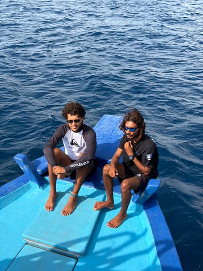 Two men sit on the blue bow of a boat on the ocean near Guraidhoo, Maldives. The man on the left has curly hair, a white, long-sleeved shirt with grey arms, dark sunglasses, and an ankle bracelet. The man on the right has long hair, a black shirt, dark sunglasses, and a watch. The ocean is blue and rippled.