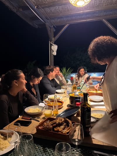 A nighttime scene depicts an outdoor dinner gathering of six adults under a thatched pergola in Casares, Spain. A large wooden table, illuminated by a woven wicker light fixture suspended from the pergola, is laden with food and beverages.

One woman, positioned at the right of the image, stands and actively serves food from a large dark pot using a ladle, into a white patterned bowl containing couscous. She has curly brown hair and wears a light-colored top, with sunglasses hanging from her neckline.

Around the table, five other individuals are seated. From left to right:
1.  A woman with dark hair pulled back, wearing a dark top, looks towards the serving woman with her left hand supporting her chin.
2.  A woman with dark hair, possibly secured with a clip, wearing a dark top, looks down at the table.
3.  A man with short dark hair, wearing a dark hoodie, sits with arms crossed on the table, observing the serving woman.
4.  A man with light brown hair, wearing a light-colored shirt, looks to his right, away from the immediate serving area.
5.  A woman with long dark hair, wearing a light-colored top, focuses intently on the serving process.

The table features multiple dishes, including a large rectangular ceramic pan filled with roasted lamb chops, several bowls of couscous, and smaller ceramic bowls containing a reddish stew or sauce. Beverages include a dark bottle of red wine (Pata Negra Valdepeñas), a bottle of olive oil, a bottle of mineral water (Fons Salutis), a beer bottle, and a glass of orange juice. Various wine glasses, some empty and some containing drinks, are arranged on the table alongside plates, cutlery, and a black smartphone.

The background is dark, indicating night. Green foliage, possibly a palm-like plant, is dimly visible behind the seated individuals on the right. A white wall or structure is also present in the distance. The pergola structure consists of dark wooden beams, with a section of rope or a pulley system attached to one vertical support on the far right.