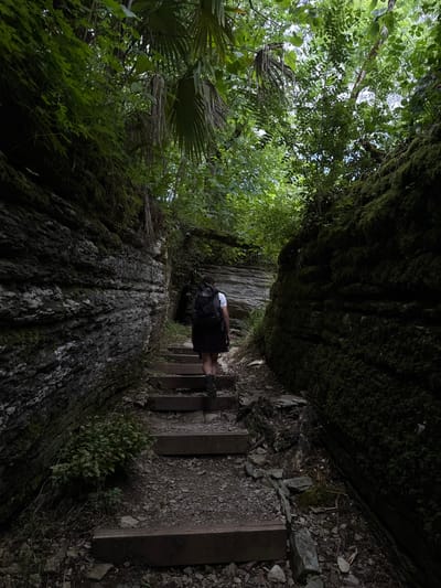 The image captures a person walking up a wooden staircase along a narrow, moss-covered path in a dense, lush forest. The path is flanked by large rocks and overhanging trees, with sunlight dappling through the canopy. The person, seen from behind, is wearing a backpack and appears to be hiking. The scene suggests a secluded and natural environment, possibly a park or a hiking trail in a remote area. The lighting and shadows indicate it could be daytime, perhaps mid-morning or afternoon, with overcast conditions that soften the light. The overall mood is one of adventure and exploration amidst nature. There is no visible text in the image.
