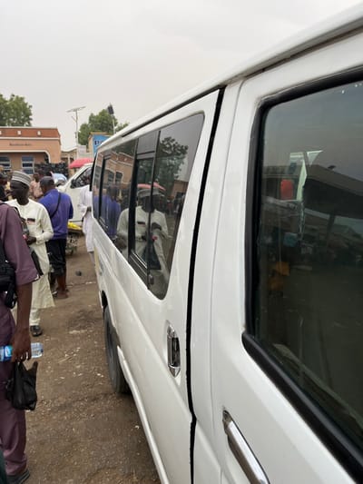 The image displays an outdoor scene in Kasuwan Kaji, Nigeria, under an overcast sky. A white passenger van is centrally positioned, viewed from its right side, slightly angled forward. The van features multiple dark-tinted windows along its side, a visible sliding door with a chrome handle, and a front passenger door also with a chrome handle. Reflections in the van's windows show portions of the surrounding environment, including buildings and individuals.
The ground surface is unpaved, consisting of packed dirt. Several individuals are visible around the van. In the immediate foreground on the left, a person wearing a dark maroon garment holds a clear plastic water bottle in their left hand and a small black bag in their right hand. To their right, a man in a light-colored top and a white cap stands near the van. Other individuals are partially obscured behind him. A man wearing a white garment and a white cap, holding a red item, is clearly reflected in one of the van's windows.
In the background, multiple light-colored buildings are visible. One building displays the text "Masallacin Mata" on its facade, while another further back has "P.R.A" visible. Utility poles and some sparse trees are also present in the background. No explicit actions beyond standing are discernible among the individuals.