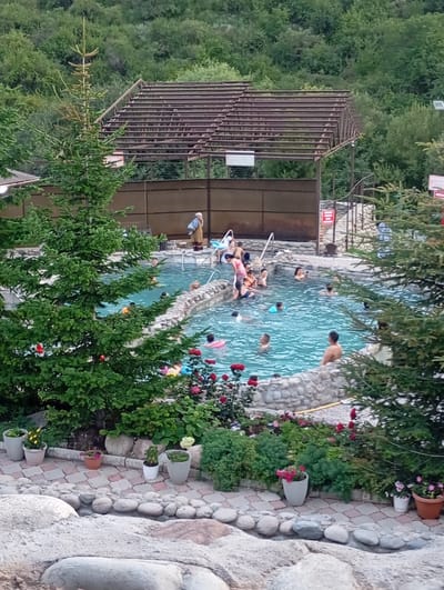 The image depicts an outdoor thermal pool, likely located in the Ak-Suu district of Kyrgyzstan. The pool, filled with clear blue water, is surrounded by a decorative stone border and lush greenery. Several individuals are visible within the pool, engaged in swimming and socializing. Some are partially submerged, while others are standing or sitting. The pool area is further enhanced by potted plants and a stone pathway leading to the water's edge. In the background, a partially constructed structure, possibly a shelter or seating area, is visible, along with dense foliage covering the surrounding hillside. The scene suggests a recreational setting designed for relaxation and enjoyment.
