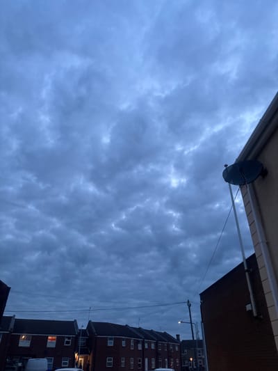 The image depicts an overcast street scene in Boston, United Kingdom, under dim, natural light. The sky is dominated by a dense covering of dark gray, lumpy stratocumulus clouds. In the right foreground, a section of a light-colored building wall is visible, featuring a dark-colored satellite dish mounted high up, with white PVC drainage pipes extending vertically downwards. On the left, a dark brick building corner is partially observed.

The midground reveals a residential street lined with two-story terraced houses constructed from dark brick, topped with dark sloped roofs. One house on the left side of the street displays a St. George's Cross flag through an illuminated window. Streetlights are active, and overhead utility poles and electrical wires are visible spanning the street. A white van is partially visible parked on the left, and another vehicle is indistinctly present further down. Two human figures are discernible near the illuminated entrance of a house on the right side of the street in the midground.
