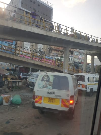An urban scene features an elevated walkway bustling with pedestrians. Below, congested traffic includes a white van with a prominent image of Jesus on the rear door, its license plate reading "KXX 449T." The setting appears to be a busy marketplace, indicated by scattered goods and informal stalls lining the roadside. The overall impression is one of vibrant urban life, juxtaposing religious imagery with the everyday realities of a crowded city.