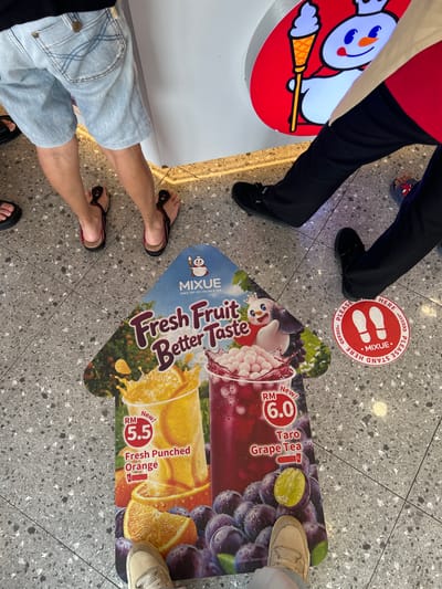 The image presents a top-down view of people's legs and feet standing on a gray speckled tiled floor, likely inside a commercial establishment. Several individuals are visible: one wearing black pants and black shoes, another in blue shorts and black and red flip-flops, and a third in light-colored pants and light-colored sneakers. A person in red attire is partially visible on the left.
The scene appears to be a waiting or queuing area, emphasized by a round red floor decal featuring white footprints and the text "PLEASE STAND HERE MIXUE". An arrow-shaped floor decal points forward, advertising Mixue's "Fresh Fruit Better Taste" beverages. It prominently displays "RM 6.0 Taro Grape Tea" and "RM 5.5 Fresh Punched Orange," both labeled "New!". A partial view of a Mixue brand standee, featuring its ice cream character, is also visible on the left side. The floor is clean and well-lit, typical of an indoor public space. The presence of Malaysian Ringgit (RM) prices on the advertisements confirms the location is in Malaysia, consistent with the provided context of Subang Jaya City Council.