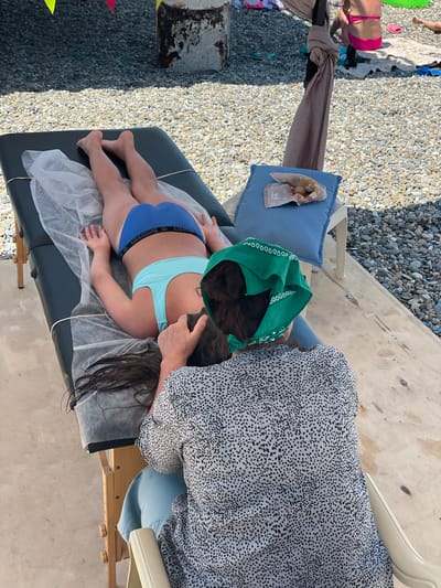 A person lies face down on a massage table covered with a white sheet. They are wearing blue bikini bottoms with the word "SUNFLOWERS" printed on the waistband and a light blue bikini top. Another person, wearing a black and white speckled shirt and a green bandana, is massaging the first person's head and neck. The massage table is positioned outdoors on a concrete surface, with a pebble-covered ground in the background. A blue cushion with a folded towel is visible to the right of the massage table. In the further background, a metal pole with draped fabric and other beachgoers are partially visible. The scene appears to be on a sunny day, likely at a beach.