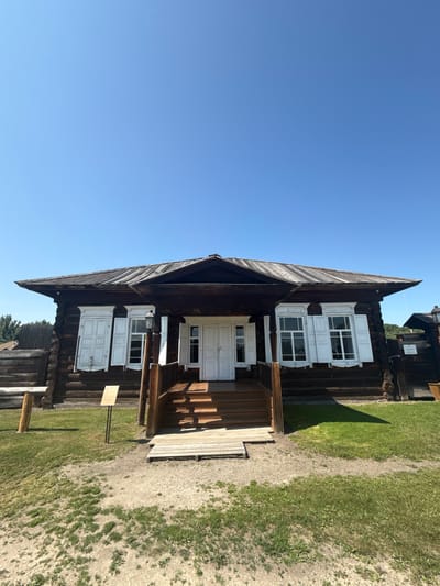 The image shows the exterior of a traditional wooden house, likely a museum exhibit, in Shushenskoye, Russia. The house has a dark wooden log construction with white shutters on the windows and white double doors leading to a porch. A wooden staircase ascends to the porch. The roof is made of weathered metal or wood. In front of the house is a grassy area with some bare patches of soil, and to the left, a wooden fence and a rustic bench are visible. A signpost stands in front of the house. The sky is clear and blue. The scene depicts a historical or cultural site, possibly related to the exile of Vladimir Lenin, as Shushenskoye is known for its Lenin memorial museum.