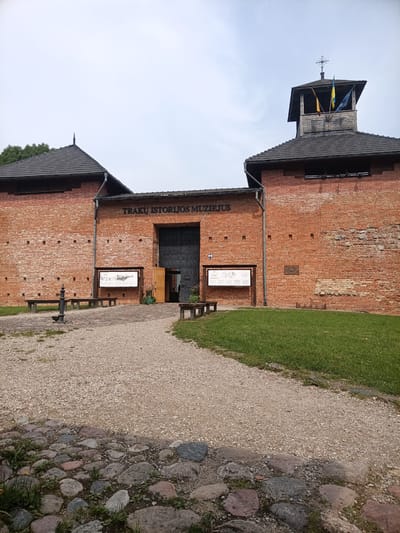 The image shows the exterior of the Trakai History Museum, located in Trakai, Lithuania. The museum is housed in a large, red brick building with a prominent tower topped with flags and a cross. The entrance to the museum is a large, dark metal gate, with an open wooden door to its right. In front of the building, there is a gravel path leading to the entrance, with a cobblestone path in the foreground. There are several benches made of wood and metal scattered around the area, as well as a vintage-style water pump. A large information board is displayed outside the entrance. The grounds are partially covered with grass and some vegetation. The sky is overcast. There are no people visible in the image.