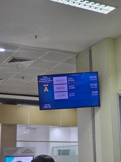 The image shows a digital flight information board at Sultan Iskandar Muda International Airport, located in Kampungblang, Indonesia. The board displays departure information for several flights.
The board is oriented horizontally and is mounted on a wall. The background of the board is dark blue, with white and yellow text and graphics.
The top left of the board displays the airport name: "Sultan Iskandar Muda International Airport by InJourney Airports". Below this, it shows "GATE A" in large yellow letters. At the bottom left, it indicates the date and time: "23 Jul 2026 06:15".
On the right side of the board, a list of flights is presented. Each entry includes the destination city, airline, and flight number. The flights listed are:
* MEDAN, IW 1213, with "GATE OPEN" at 06:45.
* JAKARTA, ID 6899, with "GATE OPEN" at 07:00.
* JAKARTA, GA 141, with "SCHEDULE" at 11:05.
The airline logos are visible next to the flight information. These include the logos for Wings Air and Batik Air. The Garuda Indonesia logo is also visible.
The environment surrounding the board appears to be an airport terminal. The ceiling is visible in the upper portion of the image, with fluorescent lighting and what appear to be air vents. The walls are painted in light yellow and cream colors. In the lower portion of the image, a glass-partitioned area and a sign that says "inJourney AIRPORTS" are partially visible. A small portion of a person's head is visible at the bottom right of the frame.