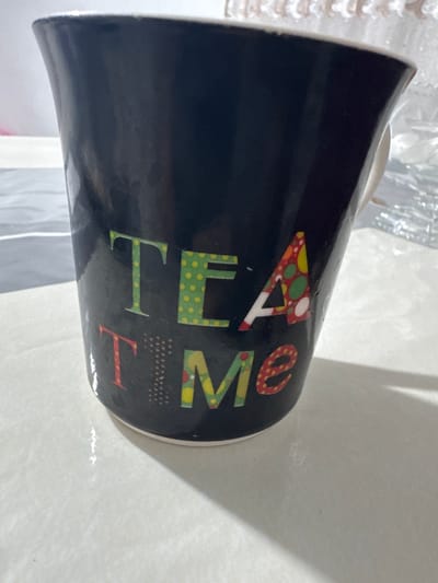 A black mug with colorful letters spelling "TEA TIME" is on a white table. The mug is angled to the right, and the lighting creates shadows on the table. The background is blurred, but appears to be indoors. The scene is a close-up of the mug. The location in the city of Ləcət, Azerbaijan is not determinable from the image.