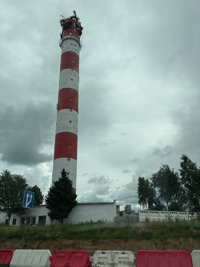 The image depicts a tall, red and white striped tower, likely a radio or communication tower, situated in the city of Lesnoy, Russia. The tower is the central focus, reaching high into a cloudy, overcast sky. It features circular windows and multiple antennas and structures at its peak. In front of the tower, a small, dark green evergreen tree stands out. A low, white building with a series of windows runs along the base of the tower, partially obscured by the tree. To the left of the tower, a blue traffic sign with white arrows indicating two lanes ahead is visible on a pole. Beyond the white building, a concrete fence with circular openings is visible, and further back, more trees can be seen. The foreground shows a dirt embankment with red and white plastic barriers. The scene appears to be captured during the daytime under a heavily clouded sky, suggesting a potentially damp or stormy weather condition. There are no people or animals visible in the image.