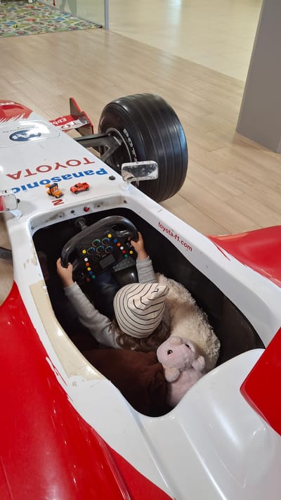 A child is seated in a red and white toy race car, holding onto the steering wheel. The steering wheel is black with colorful buttons and a digital display. The child is wearing a grey long-sleeved shirt and a striped hat. A stuffed pink pig and a fluffy white toy are also in the car with the child. The car has "TOYOTA" and "Panasonic" logos on its exterior, and "toyota-f1.com" is written on the side. The car is on a wooden floor.